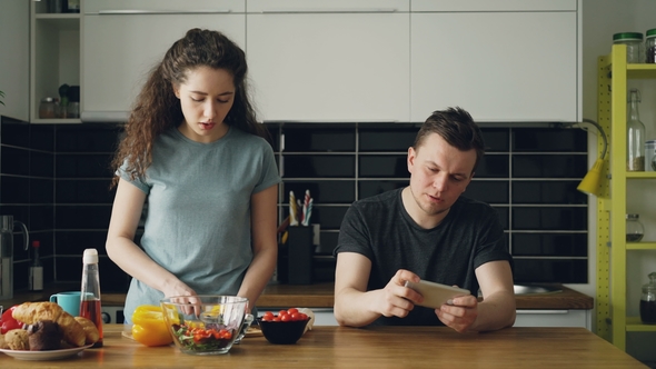 Attractive Couple in the Kitchen Man Playing Video Game on Smartphone While His Girlfriend Cooking alt