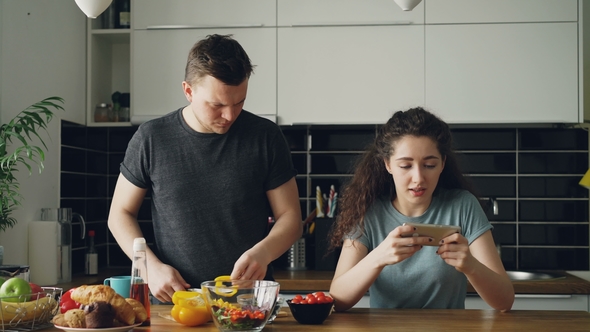 Attractive Couple in the Kitchen at Home. Curly Girl Playing Video Game on Smartphone. alt