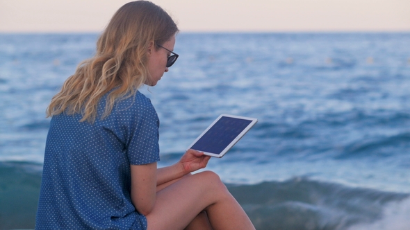 Lonely Pretty Blonde Woman Is Reading E-book By Tablet, Sitting on Sea Beach in Evening