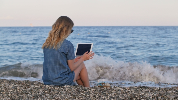 Blonde Woman Using Wifi Internet By Tablet, Sitting on Pebble Beach of Mediterranean Sea in Evening