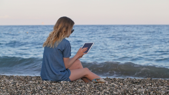 Slim Blonde Woman Looking Photo By Tablet, Sitting on Pebble Beach, Back View, Static Shot,sea Surf