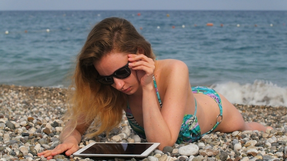 Young Blonde Woman with Long Wavy Hair Lying on Seashore, Using Tablet