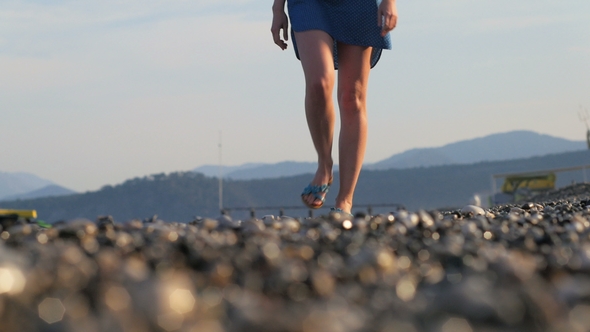 Legs of Slim Young Woman Are Walking on a Pebble Coast at Sunset, Sea Waves Are Washing Shore