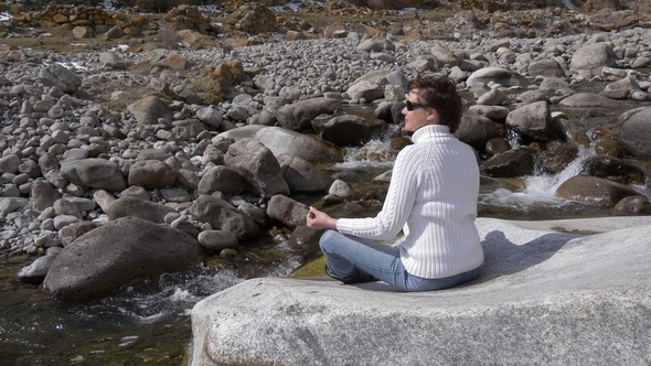 Middle-aged Woman Sitting in a Lotus Pose Near a Mountain River