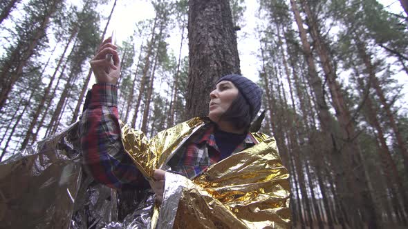 Lost Female Tourist Wrapped in a Golden Survival Blanket in the Woods Catches a Connection on Her alt