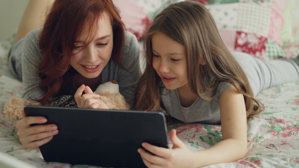Smiling Cute Girl and Young Mother in Pajamas Laughing and Looking in Digital Tablet While Lying alt
