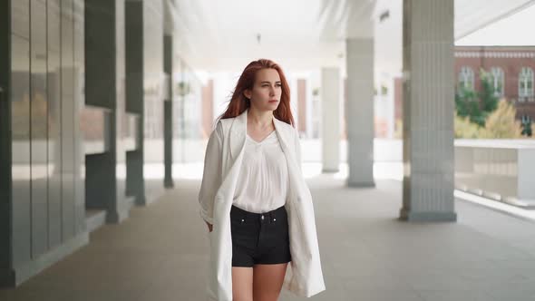 Beautiful business woman red hair in white coat stands near an office building alt