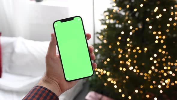 Man holding modern smartphone with green screen chromakey near Christmas tree lights on background
