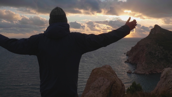 Silhouette of Man in Jacket and Cap Standing with Raised Hands and Looking at Sunset alt
