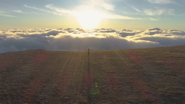 Woman Is Running Above Clouds Towards the Sun and Edge of Mountain and Rising Hands. Aerial View alt