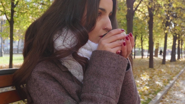 Young Southern Brunette Woman in Grey Coat Is Drinking Hot Beverage from Thermos Mug in Autumn Park alt