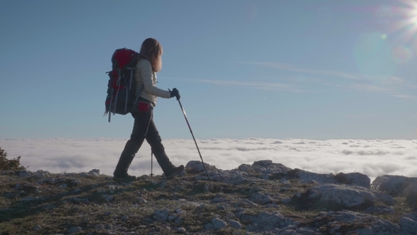 Young Woman with Backpack and Trekking Poles Is Hiking on the Edge of Mountain Above the Clouds alt