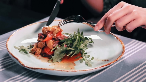 Woman's Hands with Knife and Fork Cutting Meat in Salad in Dish on Table in a Restaurant