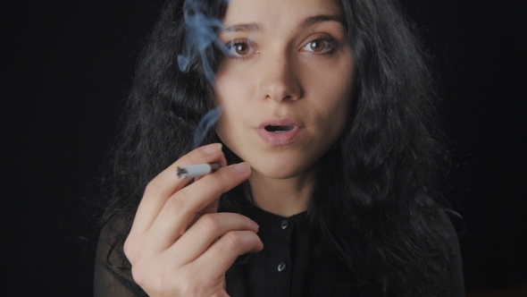 Portrait of Young Brunette Woman with Curly Hair is Smoking a Cigarette and Looking at the Camera on alt