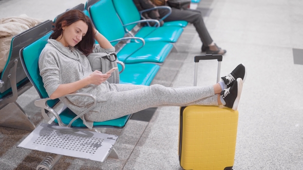 Tired Woman Is Resting in a Seat of Waiting Hall in Airport, Put Legs on Her Yellow Suitcase and alt