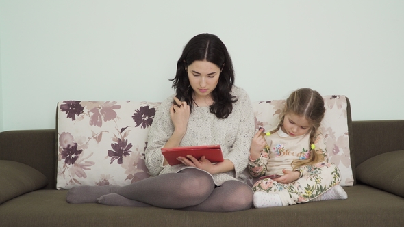 Mother Uses Digital Tablet and Her Daughter Uses Smartphone Sitting on Sofa alt