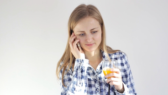 Young Woman Drinks Glass of Orange Juice Talking on Smartphone