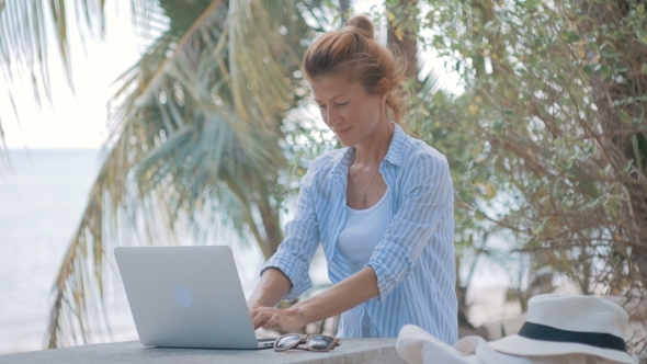 Young Woman Sitting at the Table with a Laptop with Sea View Behind alt