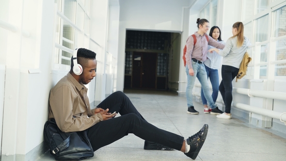 Young Handsome African American Student Sitting on Floor in White Corridor with Headphones alt