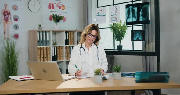Female Doctor in Glasses and Hospital Uniform Working with Paper Reports in Medical Office alt