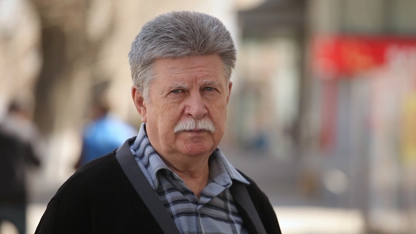 A Serious Old Man with Mustache Puts on Glasses and Stands on an Alley in Spring alt
