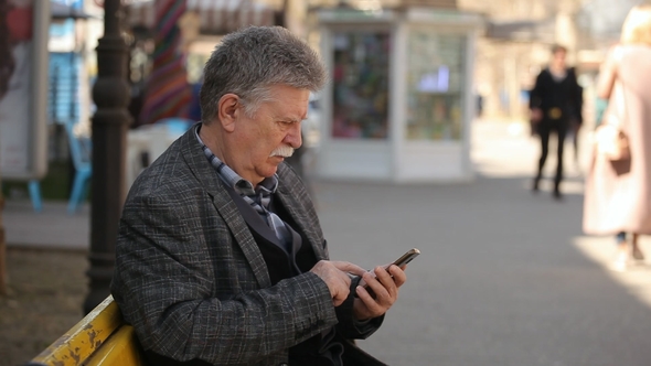 A White-headed Man Touches the Screen on His Mobile in a City Street in Spring