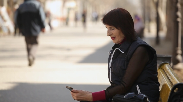 A Middle-aged Woman Logs on the Net in Her Mobile and Sits on a Bench in Spring