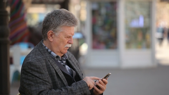 A Wise Old Man Touches the Screen on His Mobile in a City Street in Spring alt