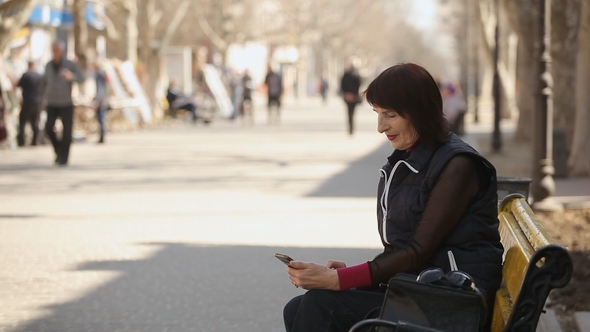 An Elegant Woman Logs on the Internet in Her Mobile and Sits on a Bench in Spring