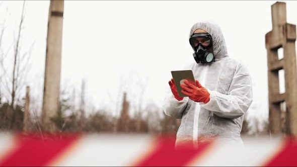 Man in Bio-hazard Suit and Gas Mask Takes Notes in His Tablet Standing on the Polluted Land alt
