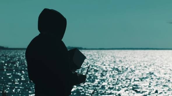 Silhouette of Man in Hood Catching Flying Quadcopter on Lake Shore