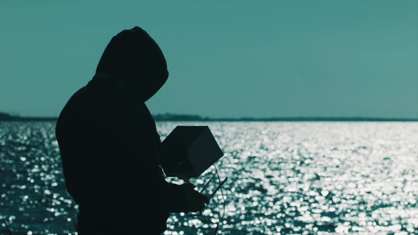 Silhouette of Man in Hood Using Drone Controller on Lake Shore