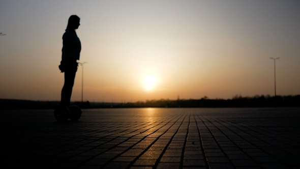 The Girl Is Driving By on the Segway Against the Backdrop of the Sunset, and a Young Man Is Walking