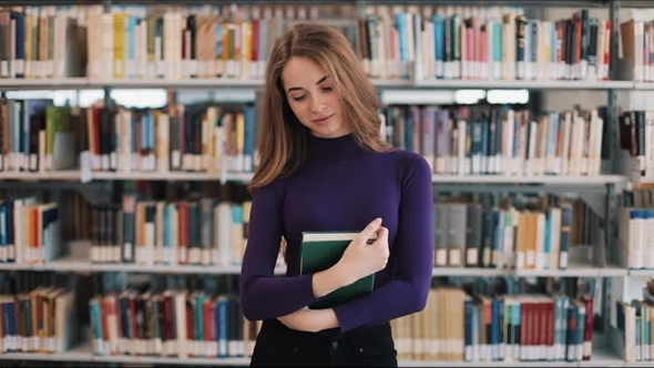 Smiling Female Student Holds a Book Standing Before the Shelves in the ...