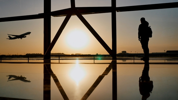 Silhouette of a Tourist Guy Watching the Take-Off of the Plane Standing at the Airport Window alt
