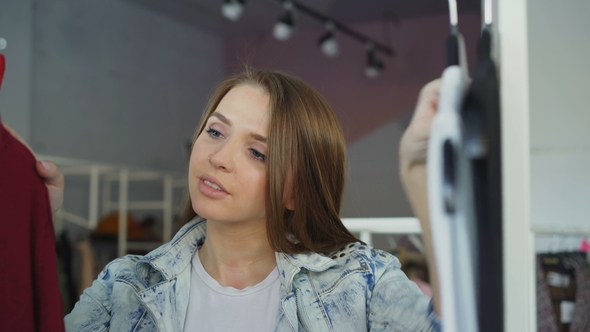 Attractive Girl Choosing Clothes in Boutique. She Is Going Through Fashionable Tops alt