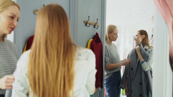 Pretty Young Woman Is Checking Fashionable Coat in Fitting Room with Her Friend Helping Her To alt