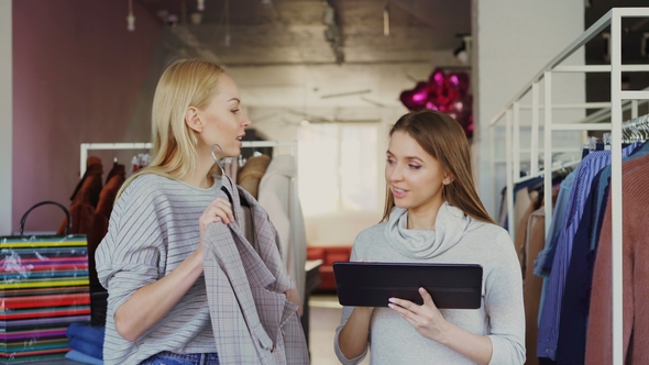 Young Business Owner Is Using Tablet While Standing in Her Clothing Store. Her Assistant Is Coming alt