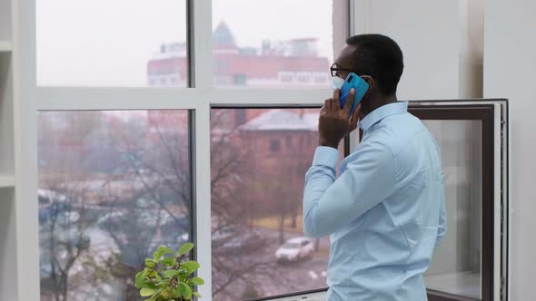 African American Male Business Man in a Medical Mask Holds a Smartphone in His Hands in an Office alt