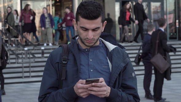 Young Middle Eastern Man Using Smartphone in City as People Are Walking Behind