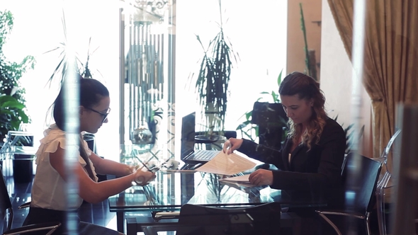 Two Business Women Discuss a Business Project Sitting at a Table in the Office Opposite Each Other alt
