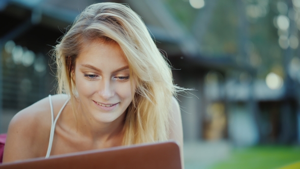 Beautiful Young Woman Enjoys a Laptop. Lies on the Lawn in the Backyard of the House in the Sun at alt