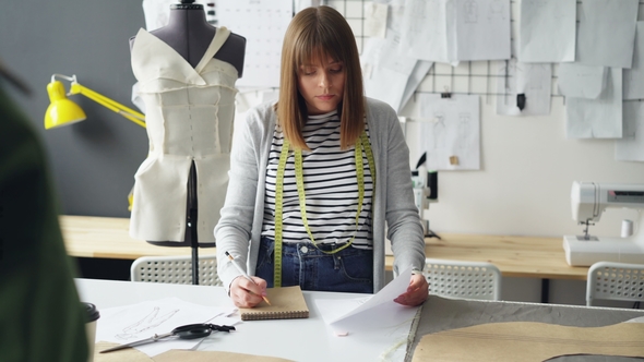 Young Woman Is Drawing in Notepad Looking at Clothing Sketch While Working in Her Tailoring Studio alt