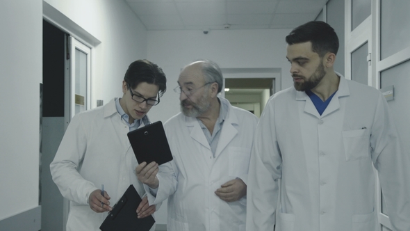 Three Male Doctors Discuss a Patient's Notes When Walk in Hospital Corridor. alt