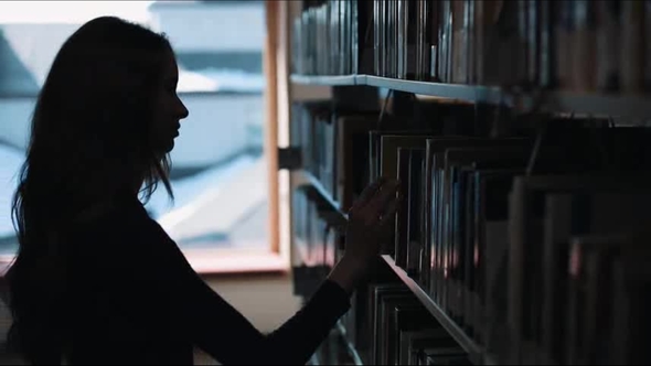 Silhouette of a Girl Looking at the Books Before a Shelf in the Library alt