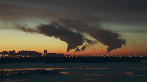 Small Town Stands Near an Icy River, White Smoke Comes From White and Red Pipes. alt