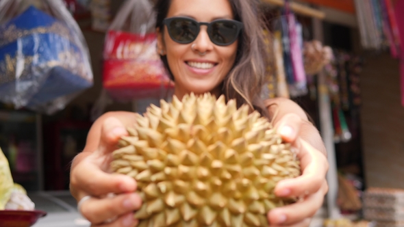 Young Attractive Smiling Tourist Girl Holding Exotic Durian Fruit in Front of Camera alt