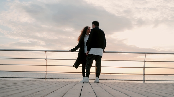 Young Happy Couple in Love. Pair Standing on Wooden Pier Near Sea or Ocean and Smiling To Each Other alt