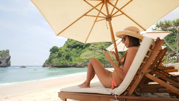 Mixed Race Young Tourist Girl Lying on Lounge Bed and Using Mobile Phone at the Beach on Tropical