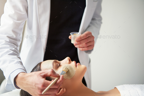 Beautician putting mask on young female face with brush - Stock Photo - Images